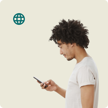 Homem jovem de perfil, com cabelo que aparenta ser afro escuro, olhando para a tela de um smartphone que segura com as duas mãos. Um ícone de globo terrestre verde-água está no canto superior esquerdo, sobre um fundo bege claro.